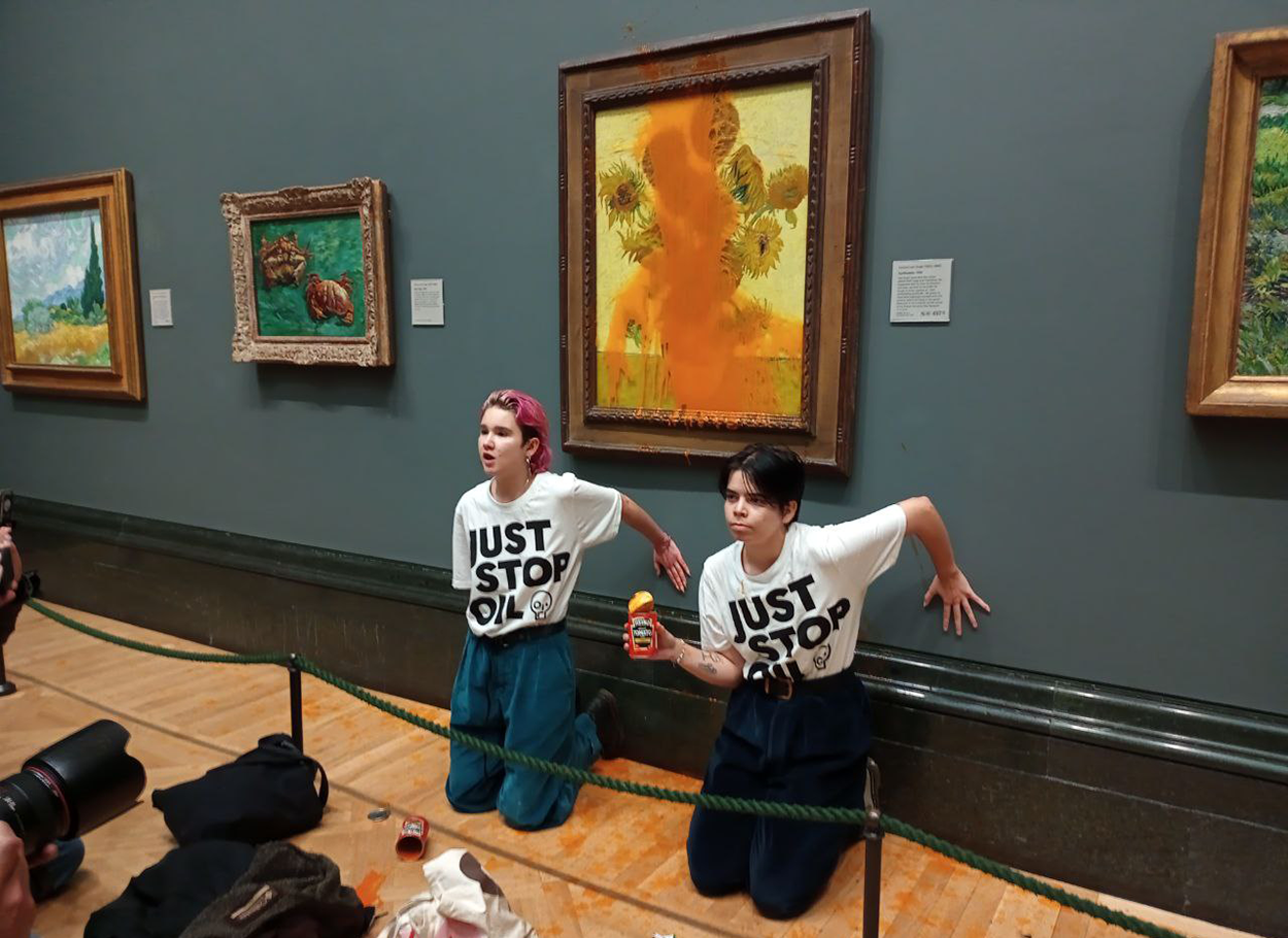 Two Just Stop Oil protesters with a soup can in front of Vincent Van Gogh's Sunflowers. They remain kneeling there with a hand on the wall and soup can in hand. Photo courtesy of Just Stop Oil. 